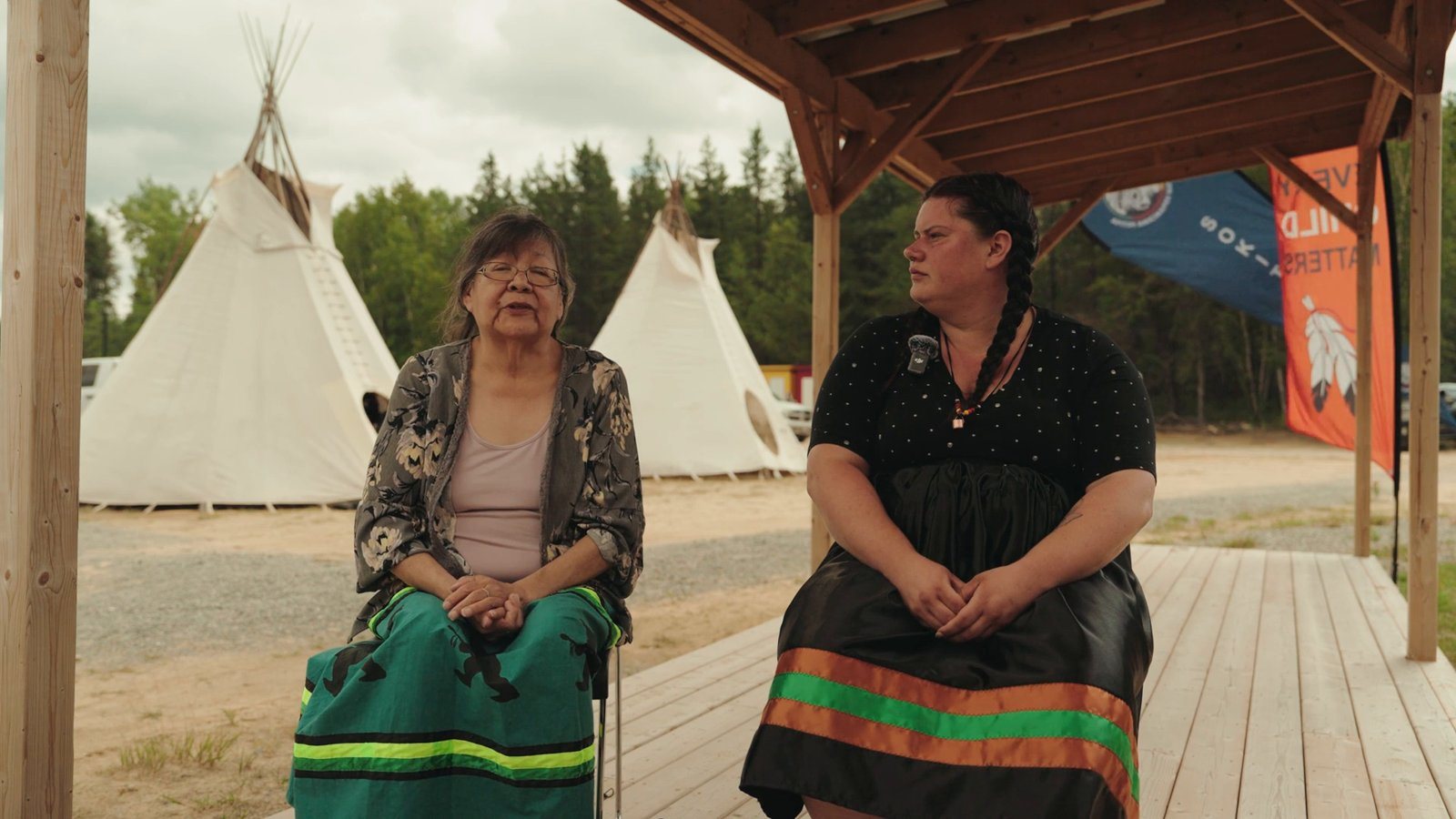 An Elder sharing her story beside a younger community member, with tipis behind them, for a Heritage Archives documentary