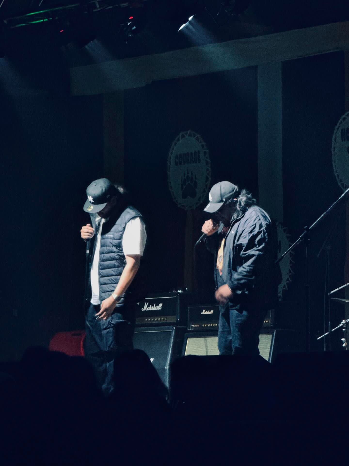 Two musicians performing on stage with microphones in hand, wearing casual outfits, highlighted by stage lights against a backdrop of amplifiers.