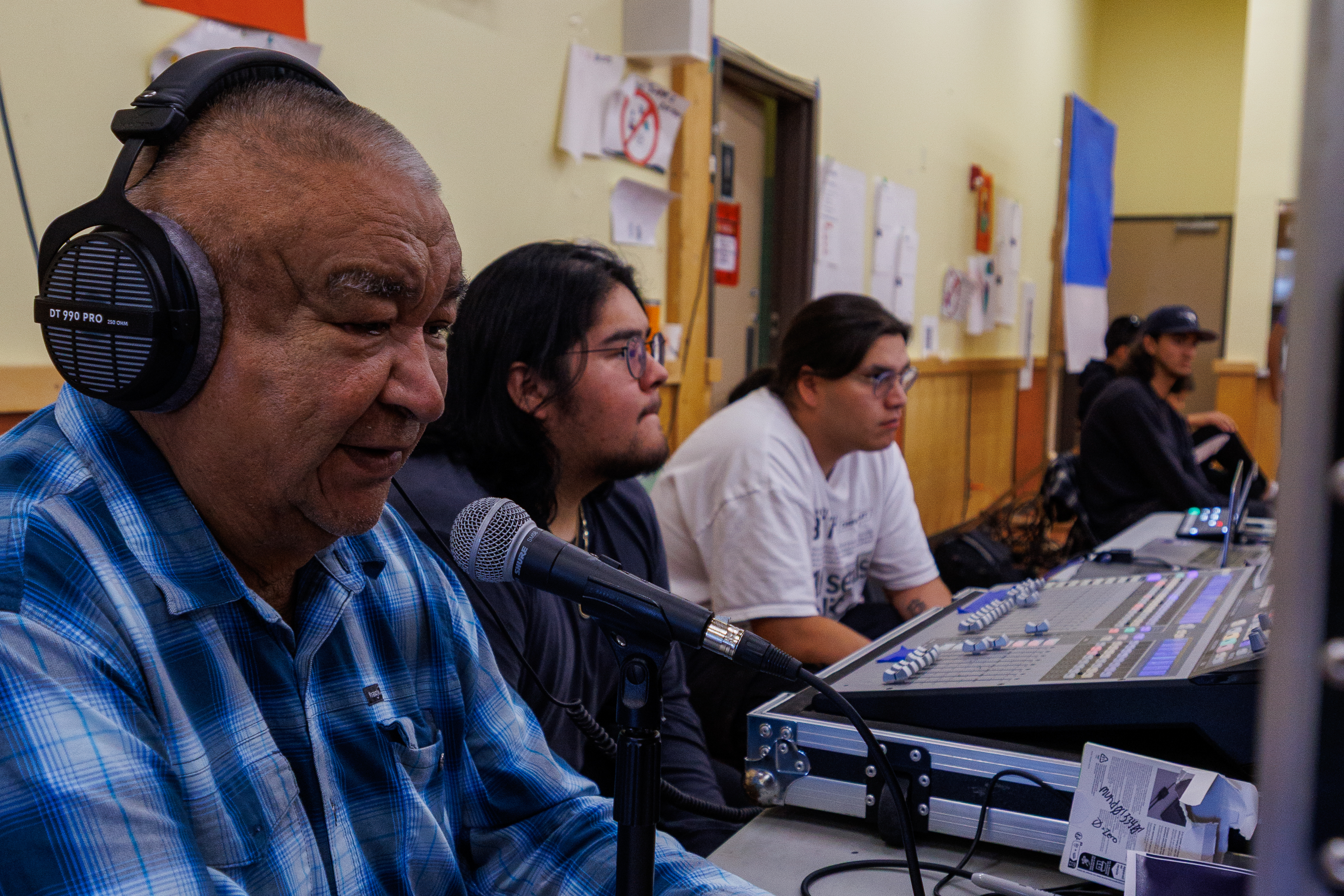 An elderly man wearing headphones speaks into a microphone while seated at a sound mixing desk, accompanied by two young men and others in a recording studio environment.