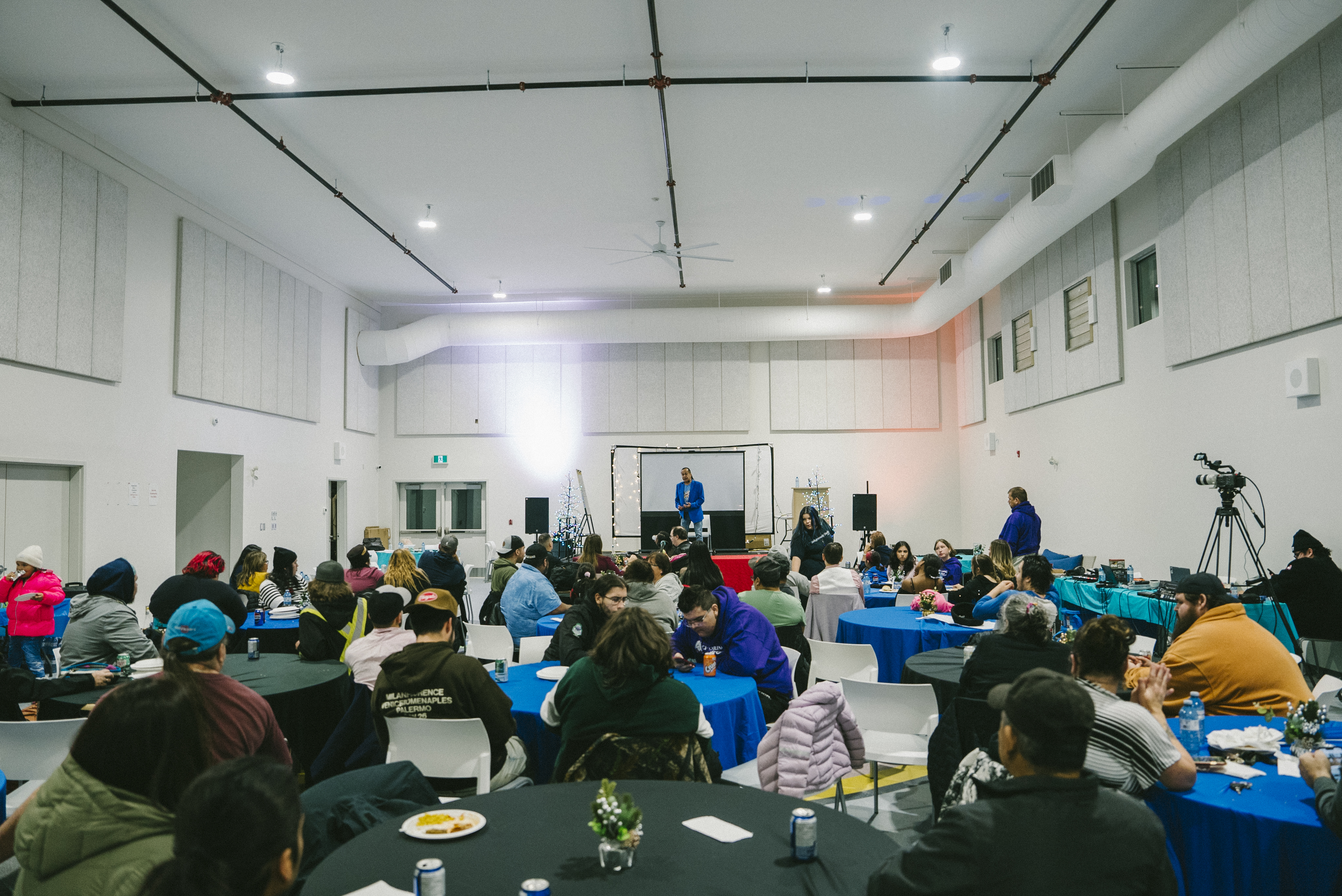 A large indoor gathering featuring several tables with people seated and enjoying food, while a speaker presents on a stage in the background. The room has a modern design with white walls and colorful table covers.