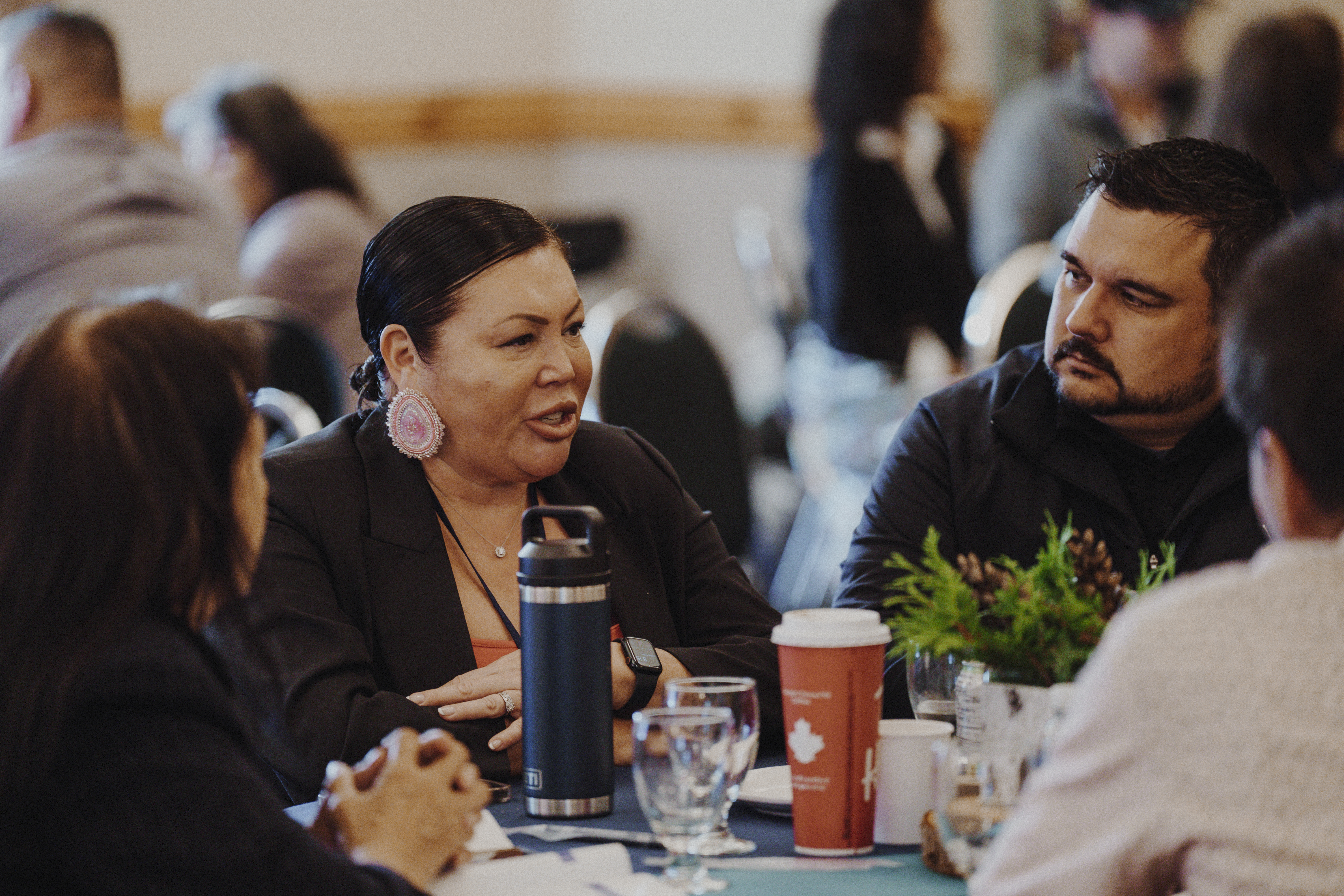 A group of people engaged in conversation around a table, featuring a woman wearing large earrings and a black blazer, a man with a beard listening attentively, and various beverages and plants on the table.