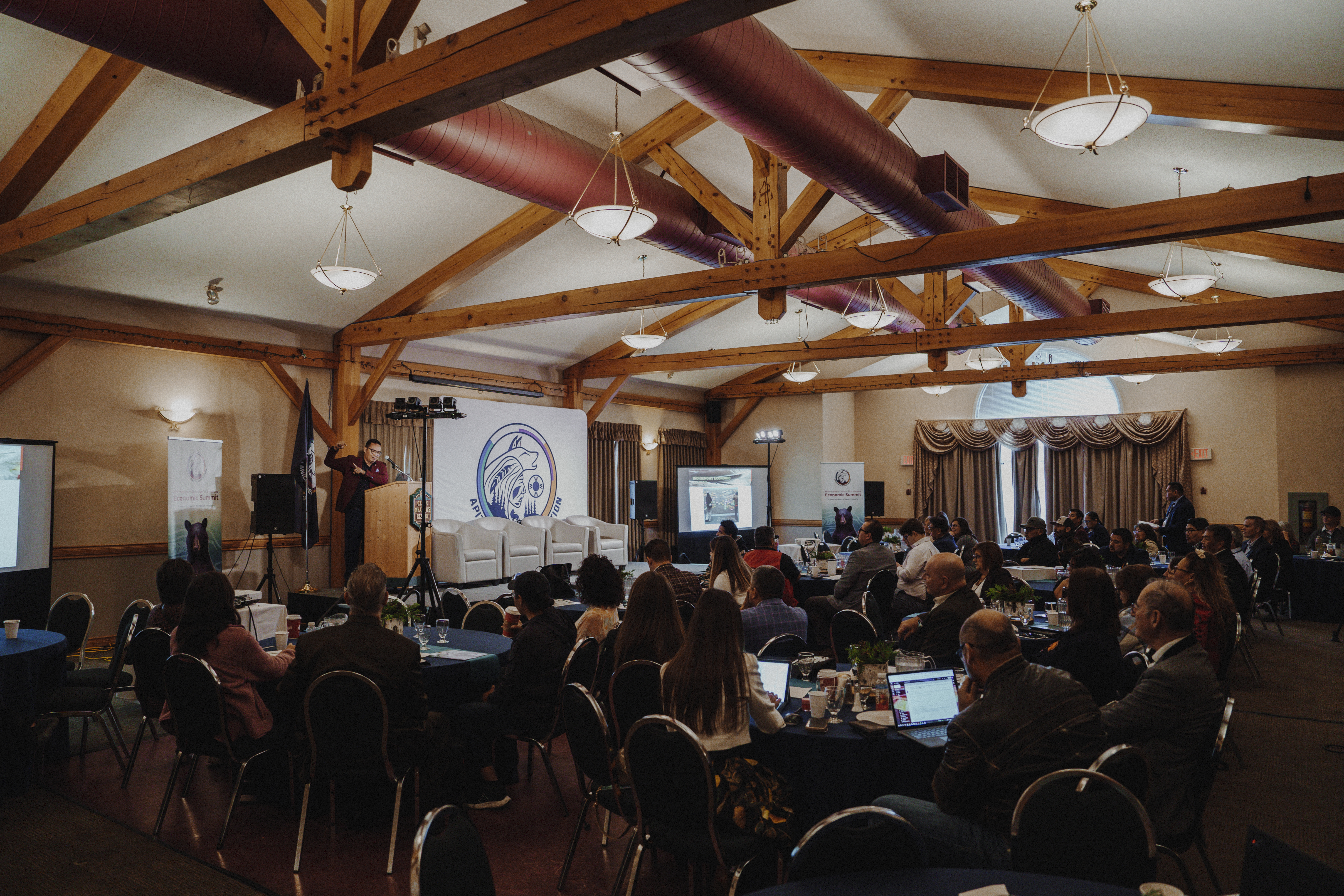 A conference room filled with attendees sitting at tables, listening to a speaker at a podium, with a projector screen displaying content in the background and wooden beams visible in the ceiling.