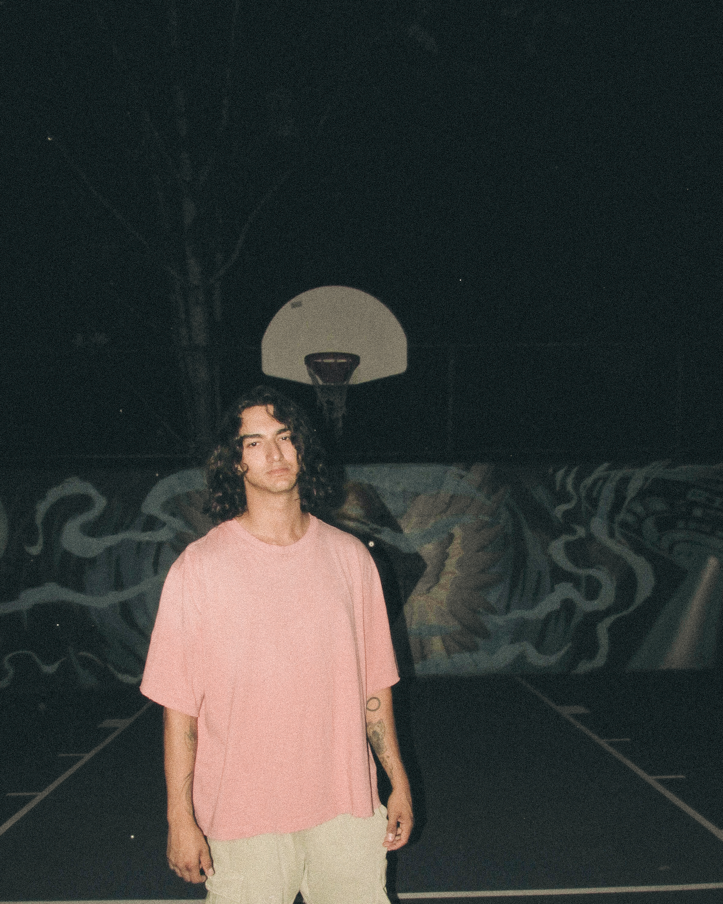 A young man with curly hair stands on a dimly lit basketball court at night, wearing a pink oversized shirt and light-colored shorts. A basketball hoop is visible in the background along with a wall mural.